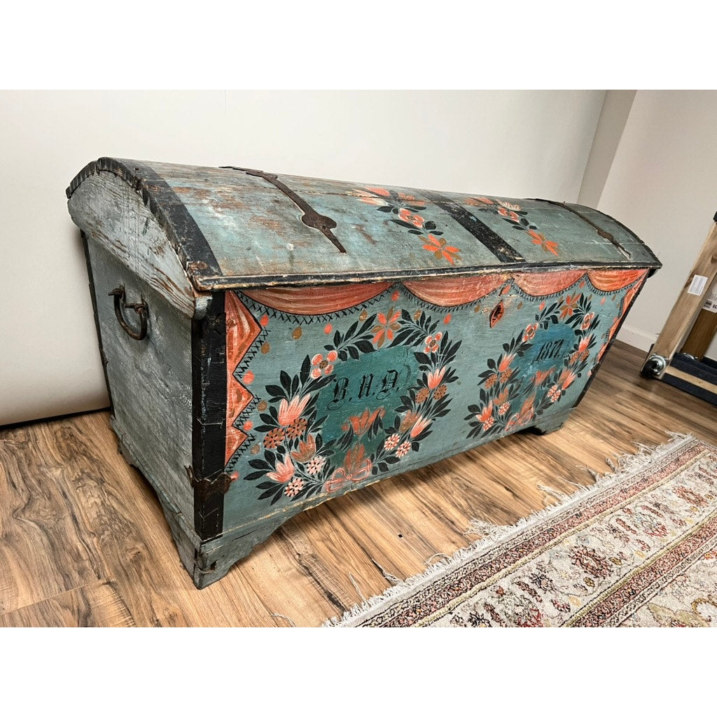 An Antique Swedish Oak Kista Dowry Chest c. 1841, with a curved lid and vibrant folk art floral motifs, stands on a wooden floor beside a patterned rug. This blue chest features metal handles and hardware. Brand: Unknown.