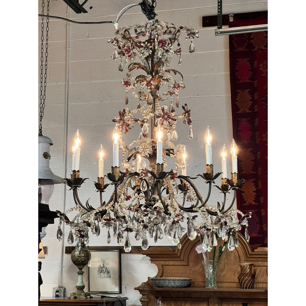 An Antique Late 19th Century French Rococo Tole Amethyst and Crystal Ten Light Chandelier hangs from the ceiling, its lit candles and crystal drops illuminating a room with framed art, a wooden mantel, and a patterned wall hanging.
