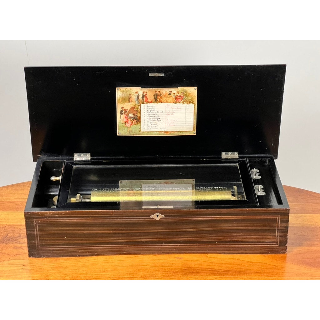 The Antique Swiss 12 Tune Music Box, crafted in the late 19th century, features a wooden case with an open lid showing its metal cylinder mechanism and a decorative label inside. Brand is unknown. The box rests on a wooden surface.