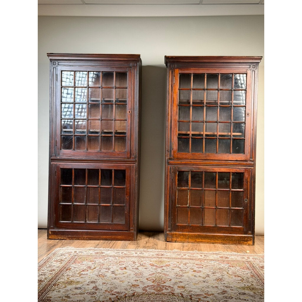 A pair of mid-19th century New England cherry glazed display cupboards (c. 1840–1860) from Pearl City Antiques stand side by side on a wooden floor against a pale wall, their glass-paneled doors evoking the charm of early American design. At the bottom of the image, an ornate floral-patterned rug is partially visible.