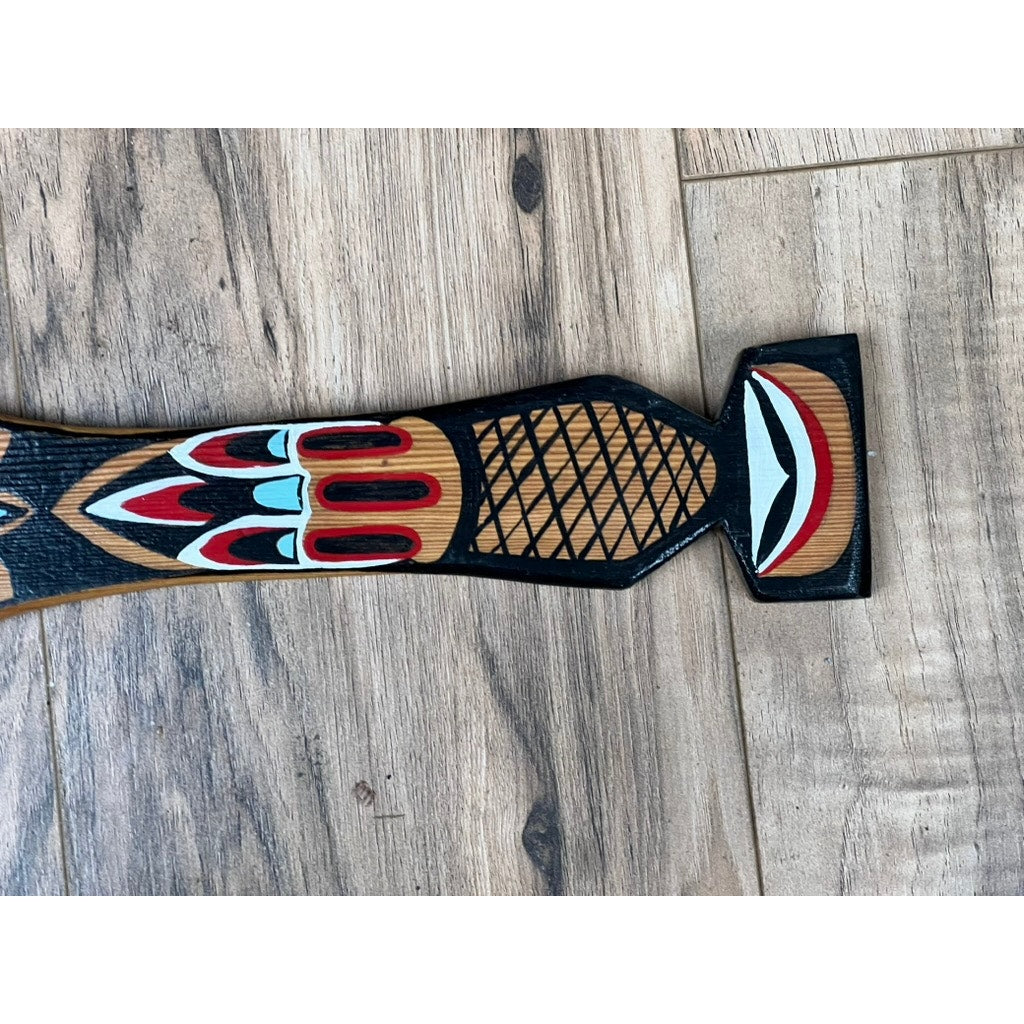 A Vintage Inuit Carved Red Cedar Paddle from Sitka, Alaska, signed illegibly, lies on a light brown wooden floor. It features intricate indigenous-style geometric patterns and bold red, black, and turquoise accents resembling a stylized animal head.