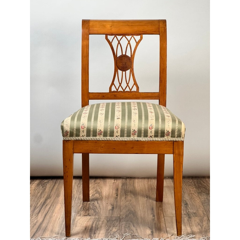 An Antique German Biedermeier Style Cherry Side Chair (early 20th c.) features a lattice and central medallion backrest, plus a green and beige striped floral cushioned seat. This elegant piece is displayed on a wooden floor.