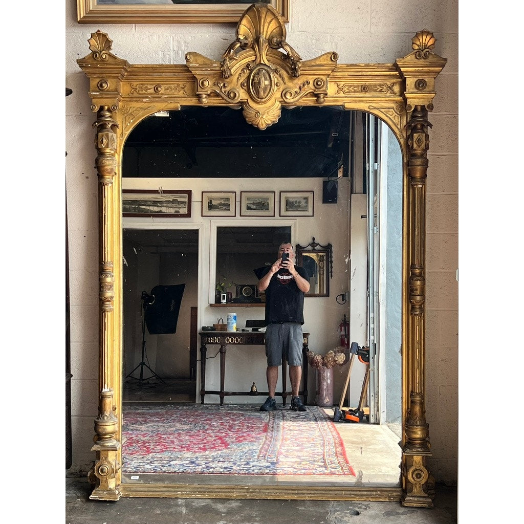 A person takes a photo in front of an Exceptional Monumental Victorian Renaissance Revival Pier Mirror, Circa 1870–1885, featuring an ornate giltwood frame, in a room decorated with art on the walls, a desk, a camera tripod, and a patterned rug.
