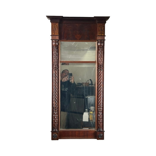 An Antique American Empire Mahogany Pier Mirror c. 1840 reflects a person taking a photo, with bottles and other items visible in the background; the mirror features an ornate carved wooden frame.