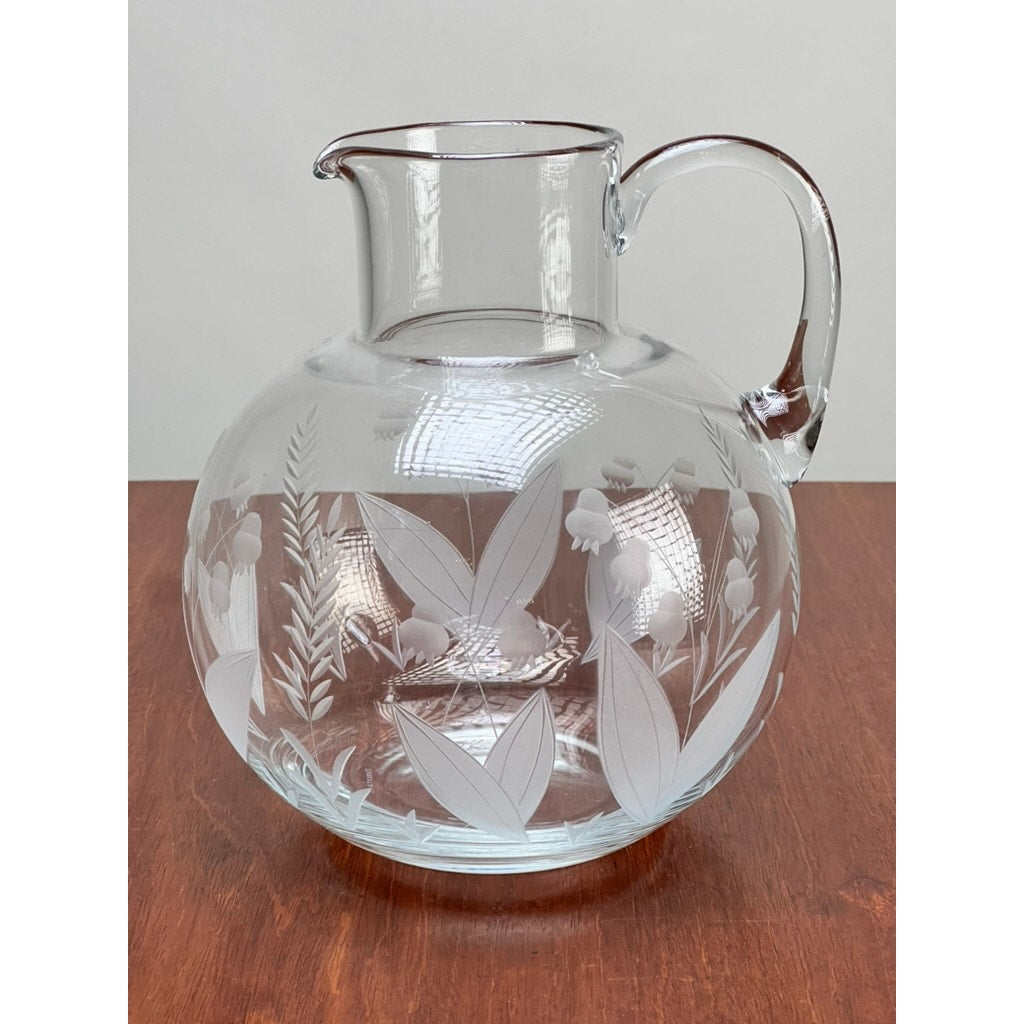 A Vintage Tiffany & Co. Etched Crystal Pitcher featuring a Lily of the Valley floral motif rests on a reddish-brown wooden surface, set against a plain light background.