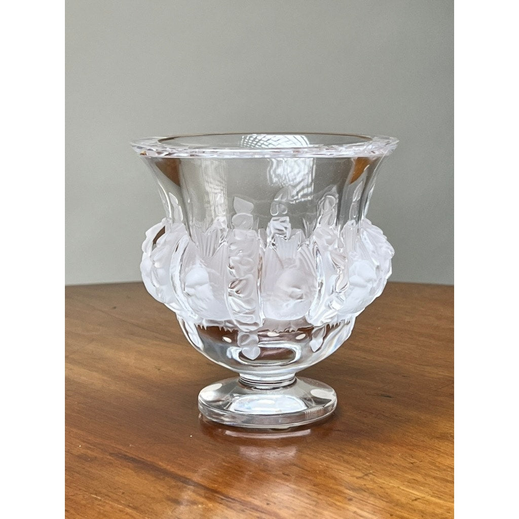 A Vintage Lalique Dampierre Sparrow Footed Vase made of clear crystal, with a flared rim and frosted leaf and berry motif by Lalique, sits on a wooden surface against a plain gray background.