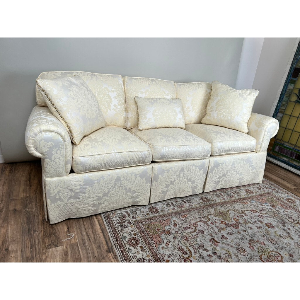 The Vintage Custom Ivory Upholstered Sofa by Stickley features three seat and back cushions plus two matching throw pillows, positioned on a wooden floor next to a patterned rug and a stained glass panel.