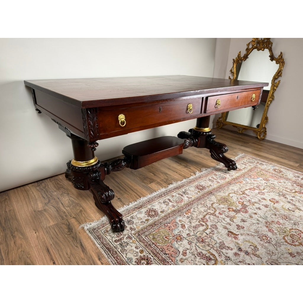 A New York Classical Revival Carved Mahogany Double-Pedestal Library Table by Ernest F. Hagen, featuring dark wood, two drawers, brass lion-head pulls, and carved pedestal bases, sitting on a patterned rug.