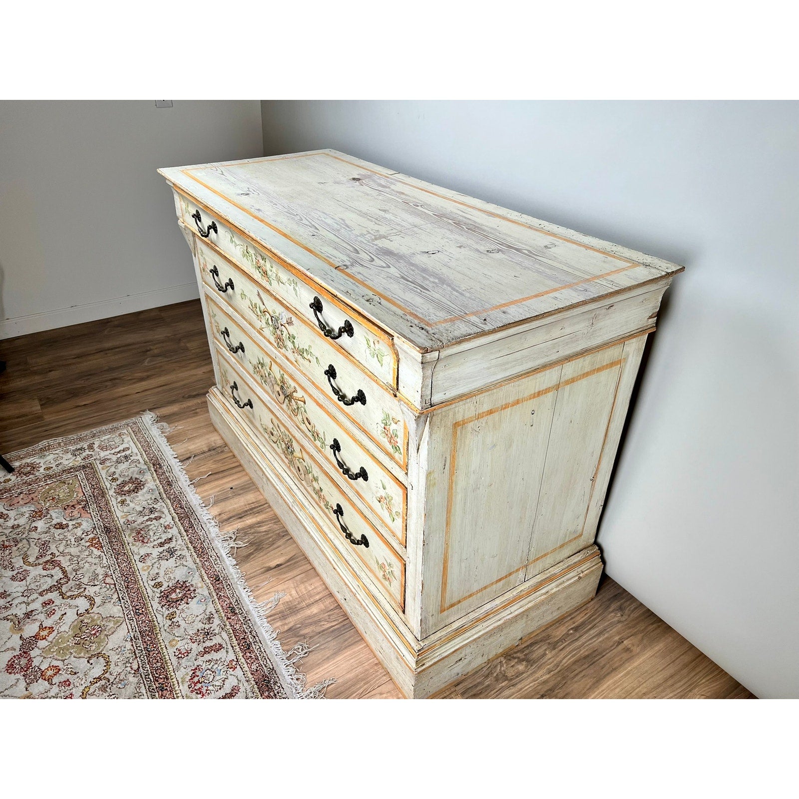 An Antique Italian Paint Decorated Four Drawer Commode (early 19th century) with ornate black handles and a distressed white finish with faded floral accents exemplifies Italian provincial style, displayed on a wooden floor near a patterned area rug.