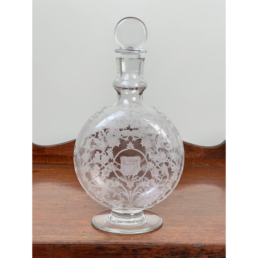 A Vintage Baccarat crystal decanter in the "Michelangelo" pattern for Martell Cognac (c. 1930) with etched floral designs and a shield motif stands on a wooden surface, showcasing intricate craftsmanship.