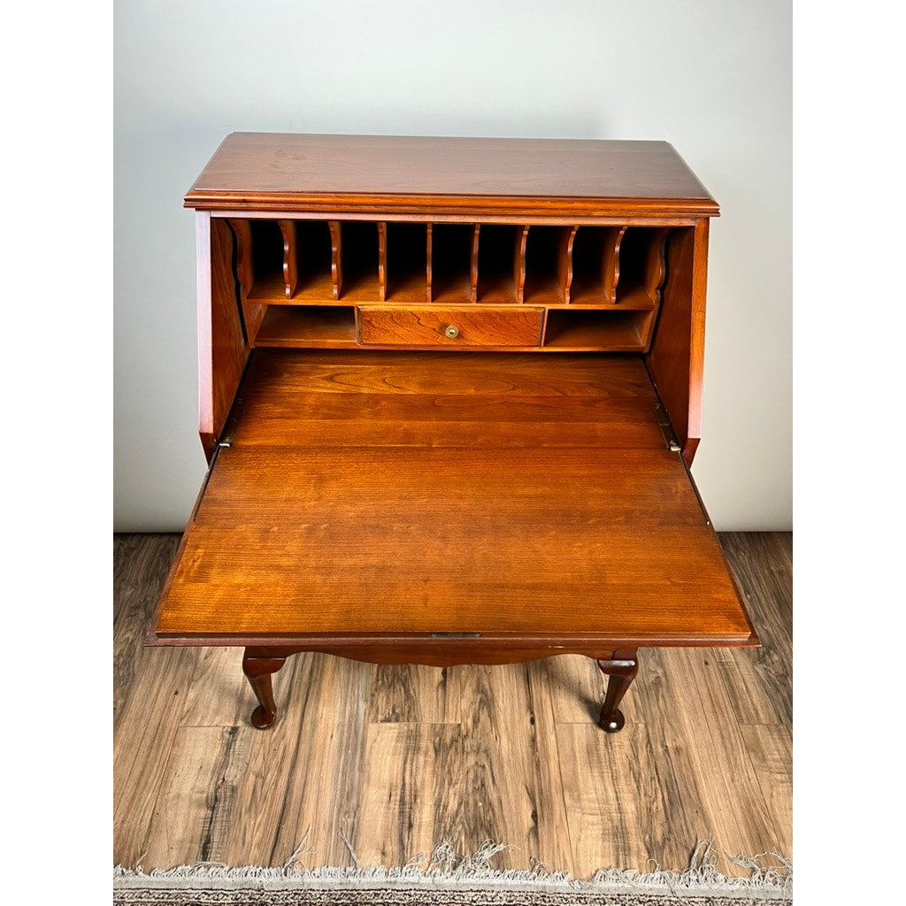 A Vintage Solid Cherry Queen Anne Style Slant Front Secretary Desk by Pennsylvania House features an open writing surface, cubby compartments, and a small drawer, standing gracefully on curved legs on a wood floor.