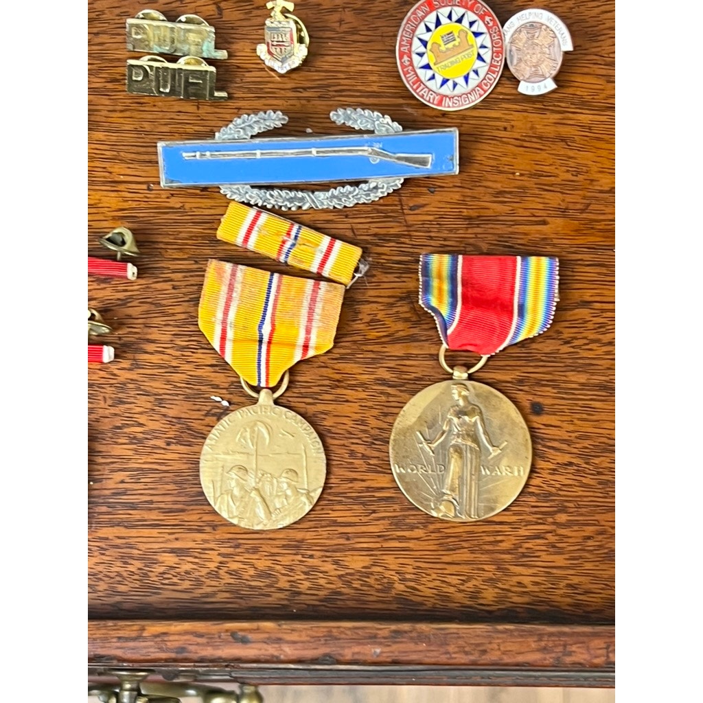 Two WWII medals with colorful ribbons from the 158th Infantry Bushmasters, attributed to Frank W. Slatcher of Company I, Pacific Theater, are shown on wood with related memorabilia—military pins and distinctive blue and multicolor circular badges.