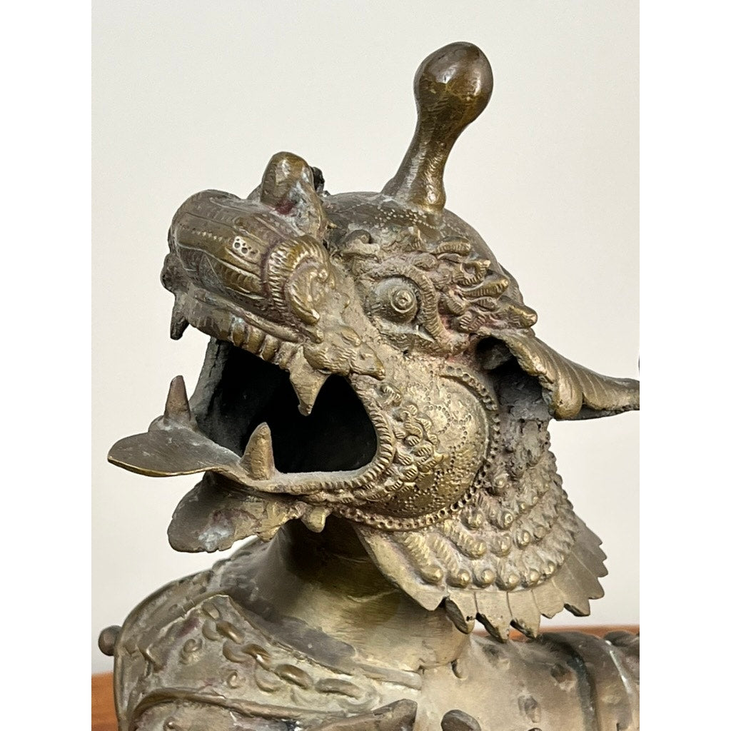 A close-up of a Vintage 17” Cast Brass Pixiu Tibetan Lion (c. 1960) shows its textured scales, ornate engravings, and decorative crest—an exquisite nod to Asian decorative arts—set against a plain background. Brand unknown.