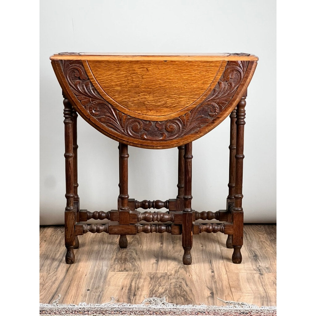 An Antique English Oak Carved Gateleg Occasional Table c. Late 19th Century, featuring ornate carved sides, turned legs, and a warm brown finish, stands on a hardwood floor against a light wall. Brand: None/Unknown.