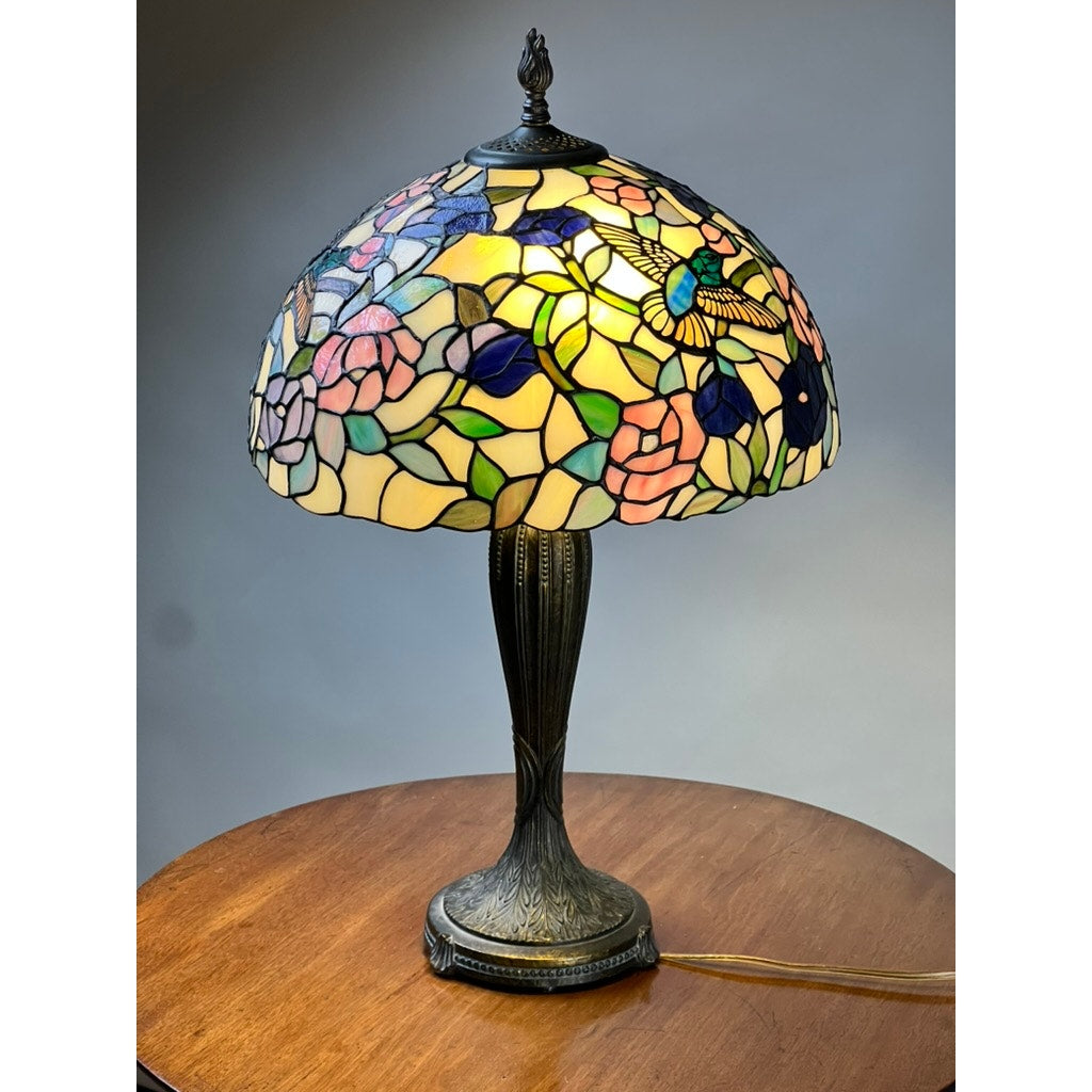 The Vintage Tiffany Style Floral Stained Glass Table Lamp features a colorful floral and hummingbird stained glass shade on a decorative Art Nouveau bronze base, perfect for adding vintage charm to any wooden surface.
