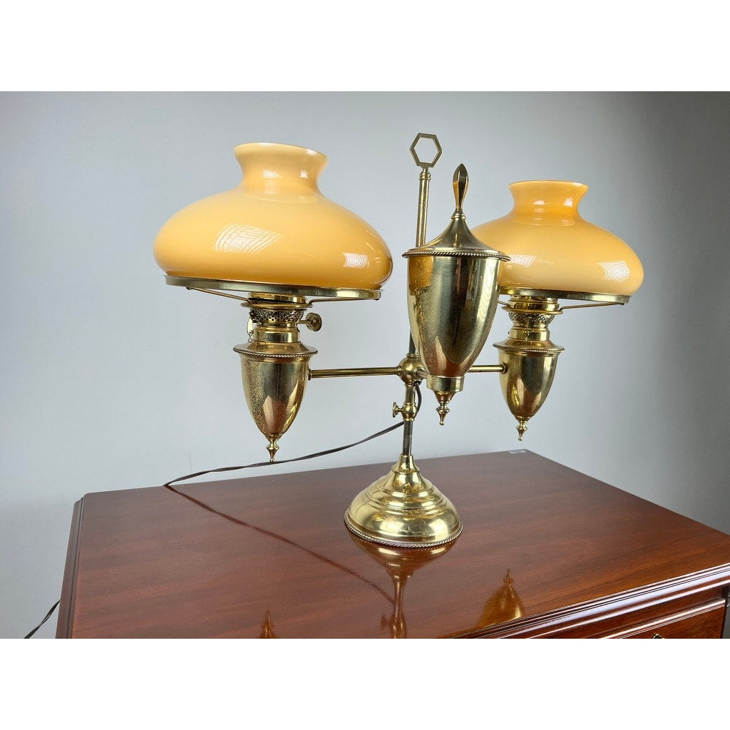A Bradley and Hubbard Antique Brass Double-arm Student Lamp with carmel shades rests on a polished wooden surface, set against a plain light gray background.