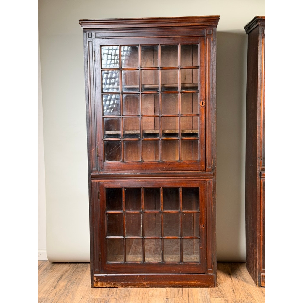 A pair of mid-19th century New England cherry glazed display cupboards by Pearl City Antiques (c. 1840–1860), featuring glass-paneled doors on both the upper and lower sections divided into square window panes, stands on a wooden floor against a neutral wall. Reminiscent of early American cupboards and glazed pieces from the era, these cabinets showcase a vintage, rustic charm that highlights their timeless appeal.