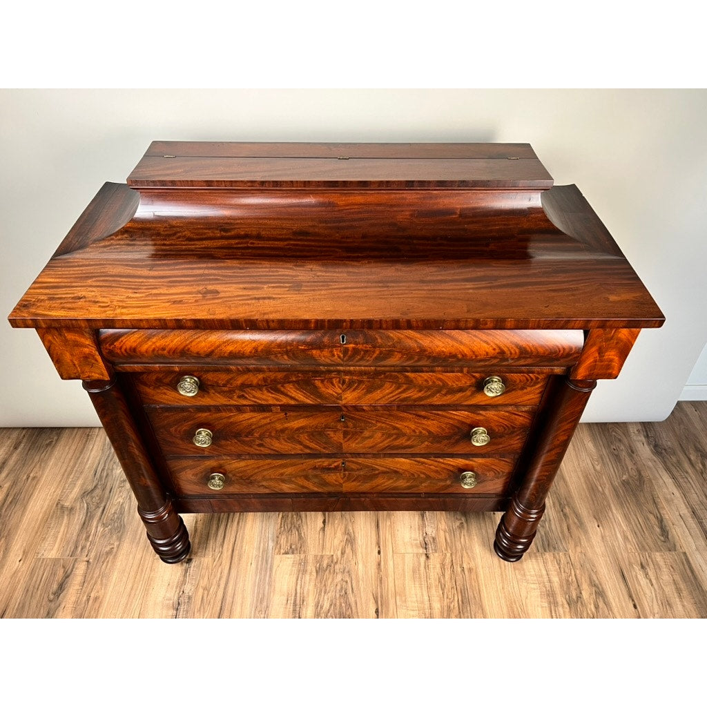 An Antique American Empire Albany Four Drawer Chest c. 1840 features flame mahogany grain, brass round handles, and carved front legs, shown on a wood floor against a plain light-colored wall. Brand: None/Unknown.