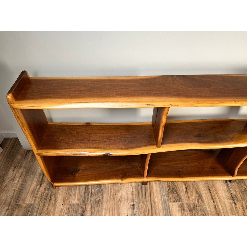 A Vintage Mid-Century Modern Style Live Edge Walnut Bookshelf with three shelves and a natural, uneven edge stands against a light-colored wall on a wooden floor.