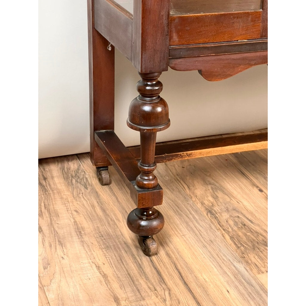 Close-up of an intricately turned wooden leg from a Depression Era Mahogany Curio Display Cabinet with Glass Sides, Circa 1930s. The leg is attached to a square frame and finished with a small caster wheel at the bottom. Resting gracefully on a light wood floor, this detail highlights the classic vintage American craftsmanship found in this elegant display cabinet.
