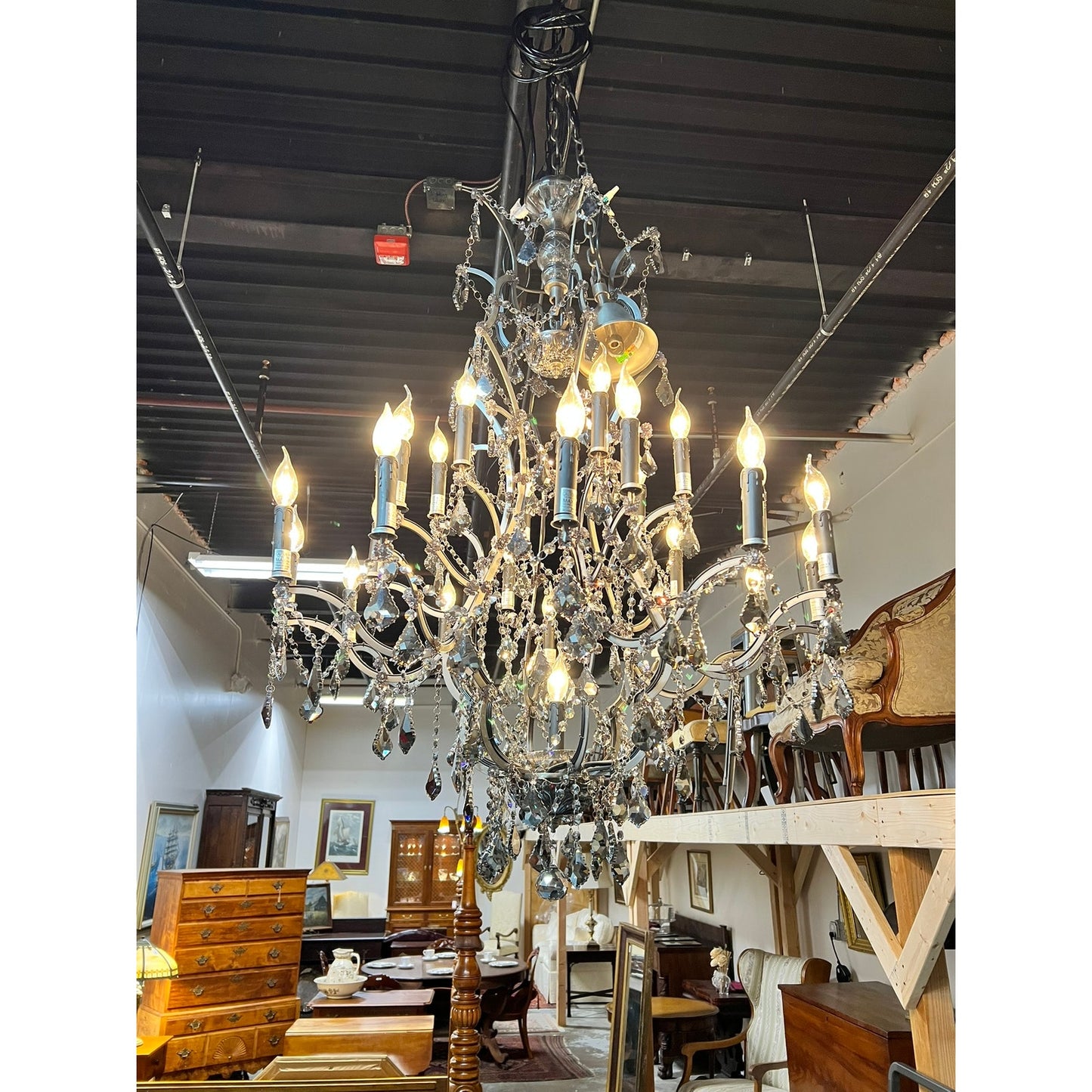 A Large Vintage Smoke Crystal 24 Light Chandelier glows from a dark ceiling in a showroom displaying wooden cabinets, tables, and other antique furniture. Brand: None/Unknown.