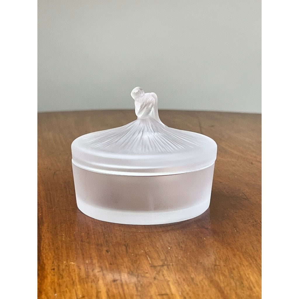A Vintage Lalique France Ballerina Frosted Crystal Covered Powder Box by Lalique, featuring a sculpted ballerina on the lid, sits gracefully on a wooden surface, evoking classic elegance.