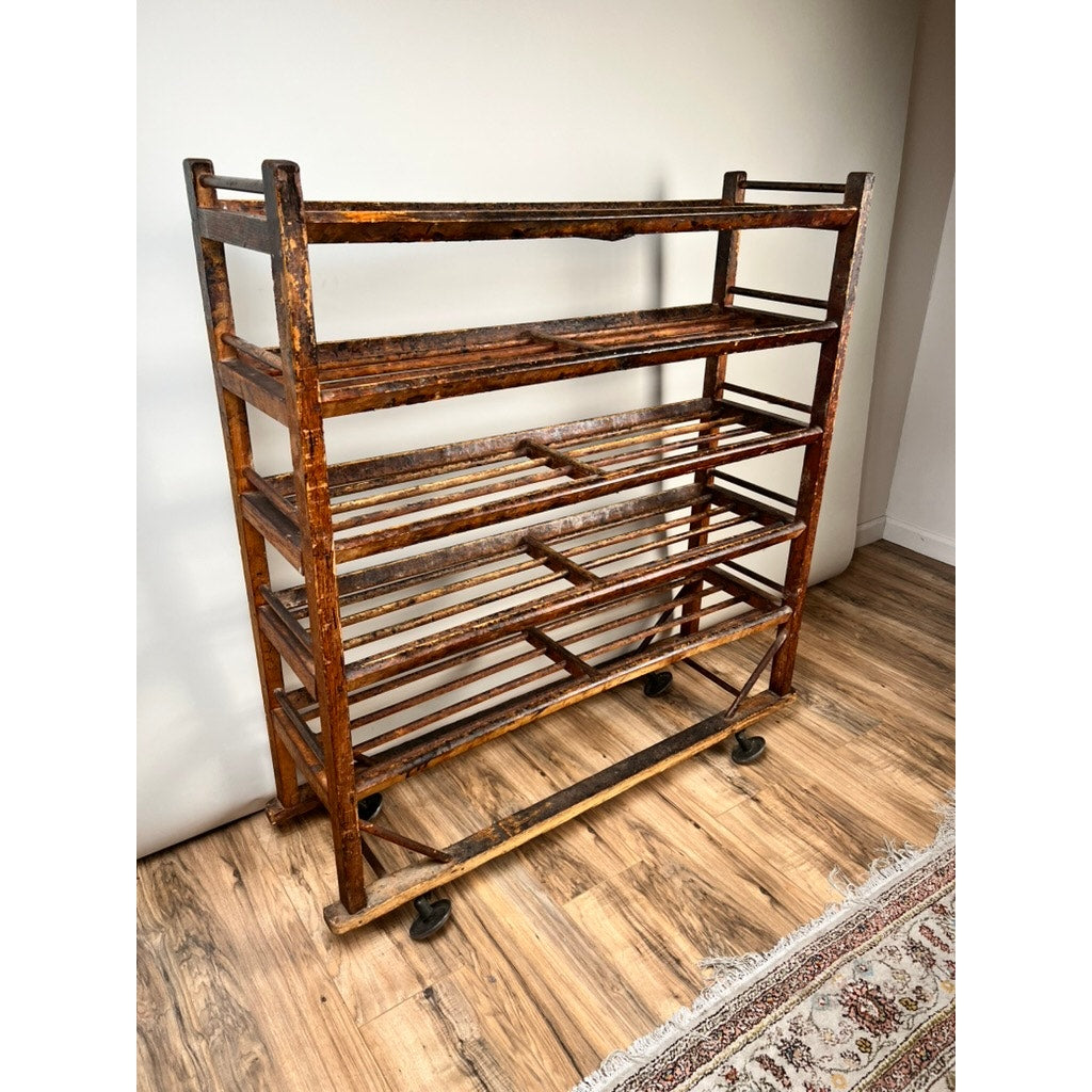 An Antique Oak Shoe Factory Rolling Rack on original industrial casters (late 19th century) stands on a wooden floor beside a patterned rug and white wall, its rustic, worn finish adding vintage character to the space.