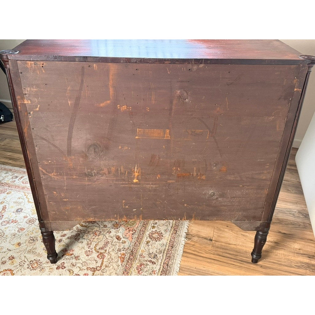 The reverse side of an Early 19th Century New England Federal Mahogany Bow-Front Chest of Drawers (c. 1810–1825), with visible scratches and marks, shown resting on a patterned rug atop a wood floor.