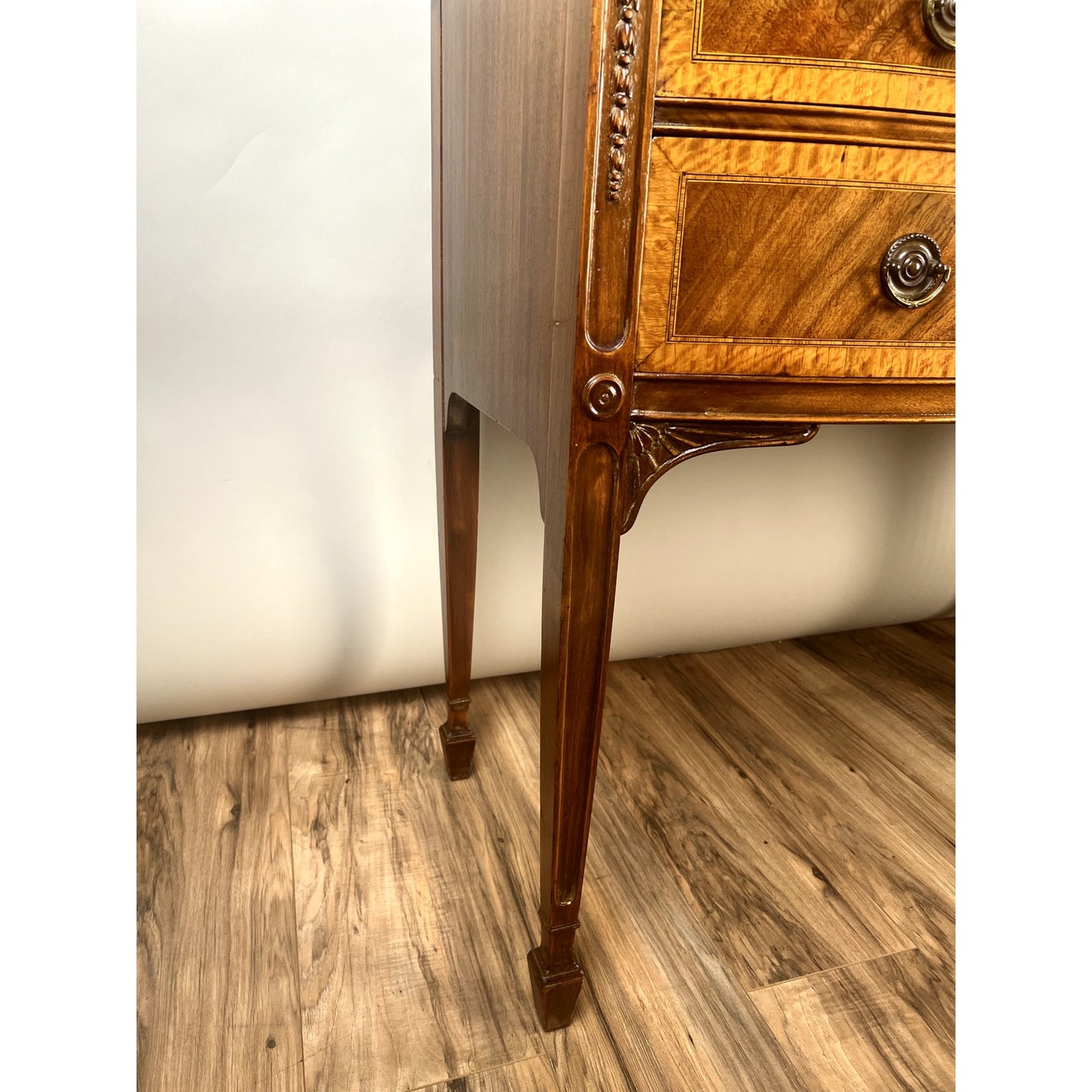 Close-up of the corner and legs of a Vintage Federal Style Bow Front Mahogany Sideboard by Paine Furniture Boston, featuring carved details and brass drawer pulls, shown on a wood-patterned floor against a light background.