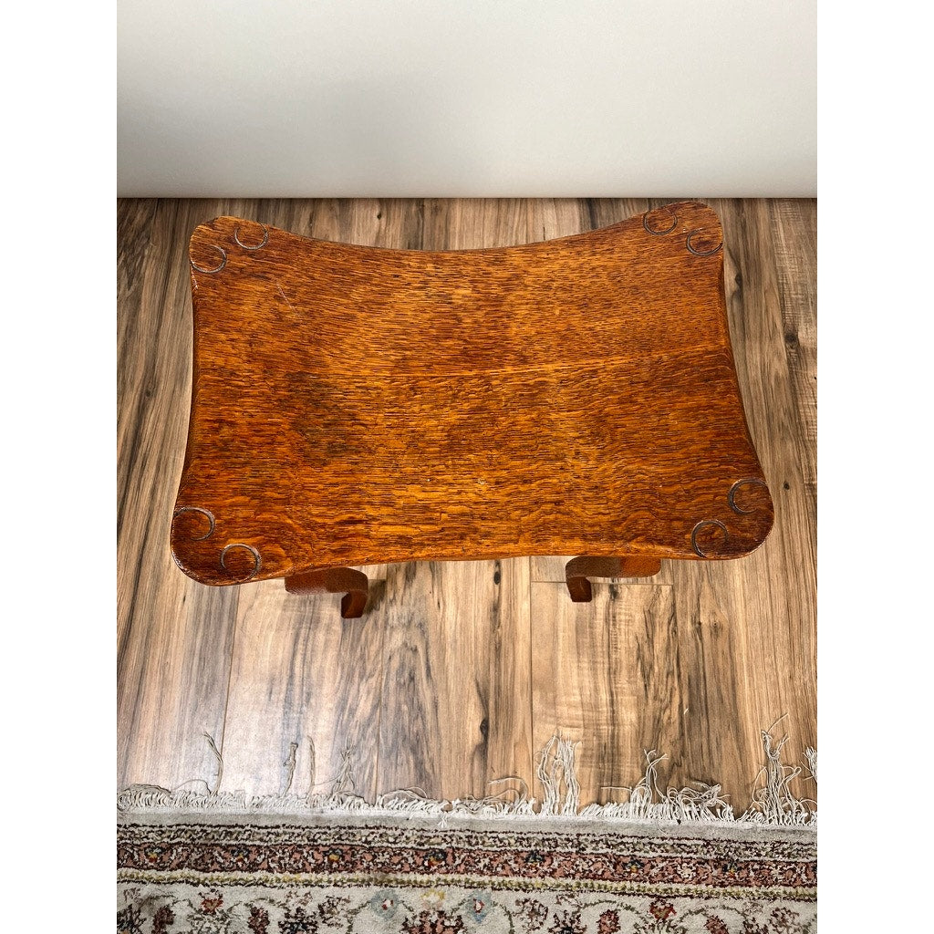 An Antique Arts and Crafts Saddle Shaped Footstool c. 1900 rests on a wood floor by a patterned rug, beside a wooden table with curved edges and decorative corner carvings. Brand: Unknown.