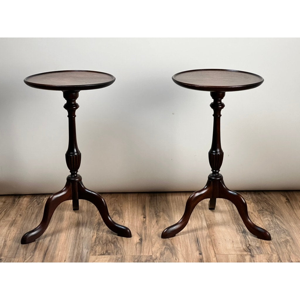 A pair of vintage diminutive walnut candle stands from circa 1940, each with a round top and three curved legs, stand on a wooden floor against a plain white wall.