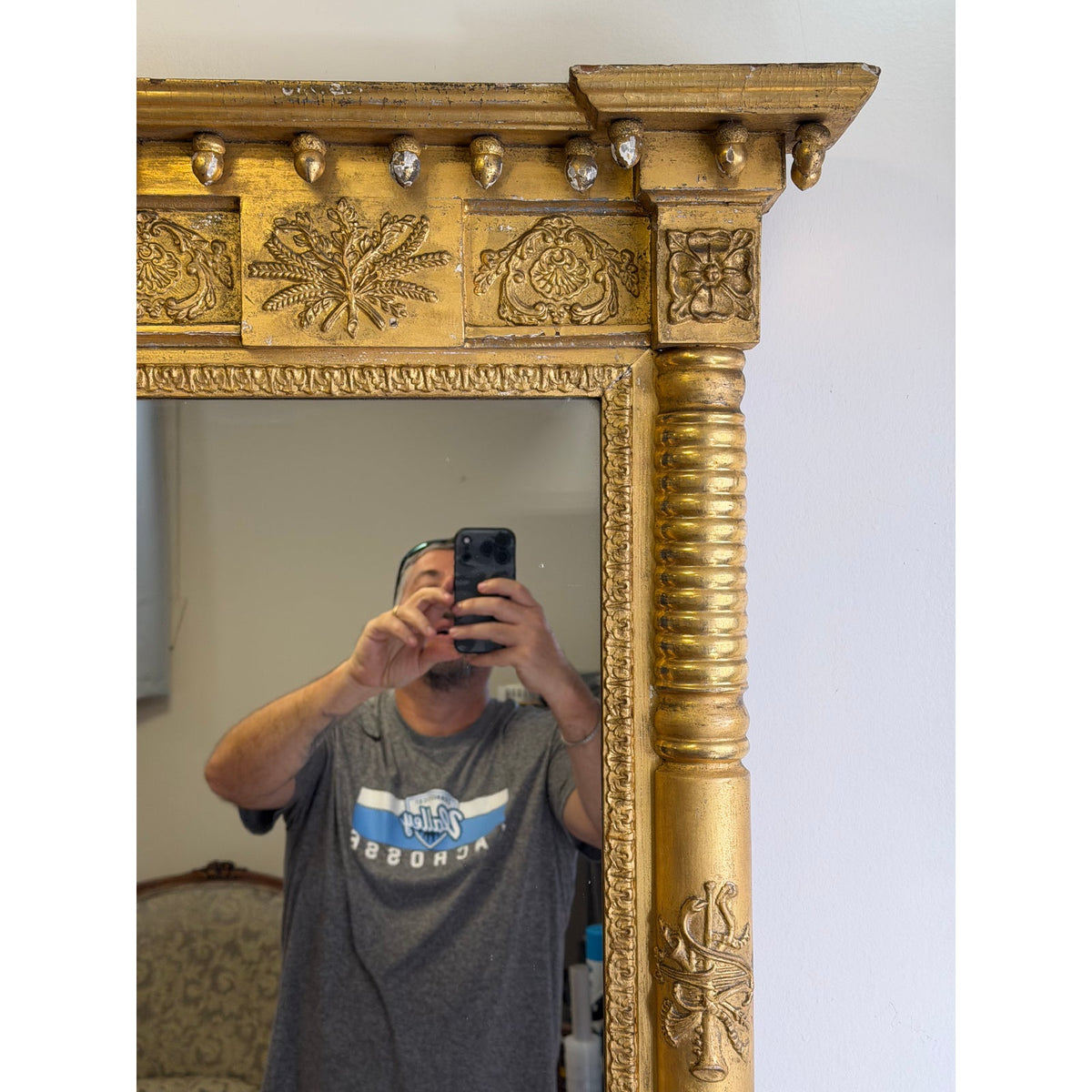 A person holds a phone to take a photo in the reflection of an American Federal Giltwood Pier Mirror, Possibly Massachusetts or New York, circa 1815–1830, featuring ornate, 19th-century classical architectural carvings and a gold-painted frame.