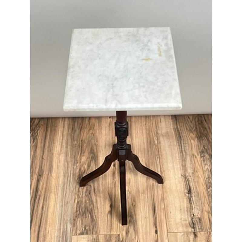An Antique Victorian Marble Top Plant Stand from the late 19th century, featuring a small square marble top, dark wooden pedestal base, and three curved legs, is set on a wood floor against a plain light-colored wall.