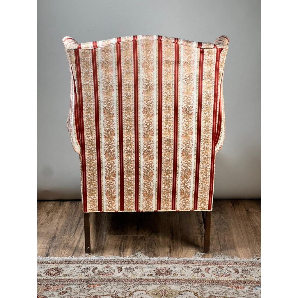 A Vintage Chippendale Style Wingback Chair by Ethan Allen, featuring red and gold traditional striped upholstery with ornate patterns, is placed on a wooden floor near a patterned rug against a plain light gray wall.