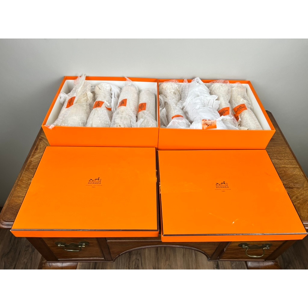 Two bright orange Hermès boxes are open on a wooden desk, displaying a set of eight Hermès Crystal Champagne Flutes by Saint Louis France (10.5”), wrapped in white tissue paper. The Hermès logo is visible on the box lids resting in front.