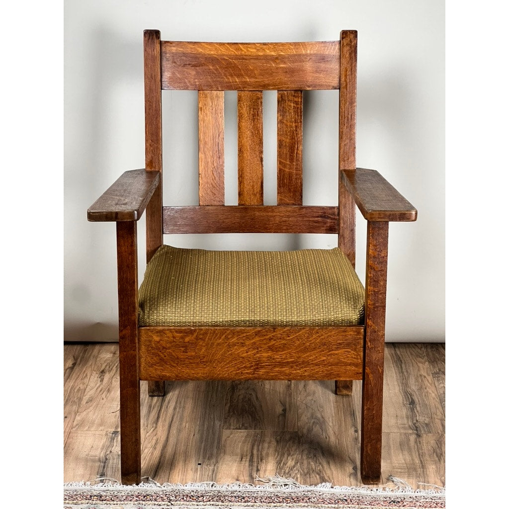 An Antique Quarter Sawn Mission Oak Arm Chair by Quaint Furniture c. 1920, with a straight back, vertical slats, wide armrests, and a green upholstered seat, sits on a wooden floor against a light wall—a true Arts & Crafts classic.