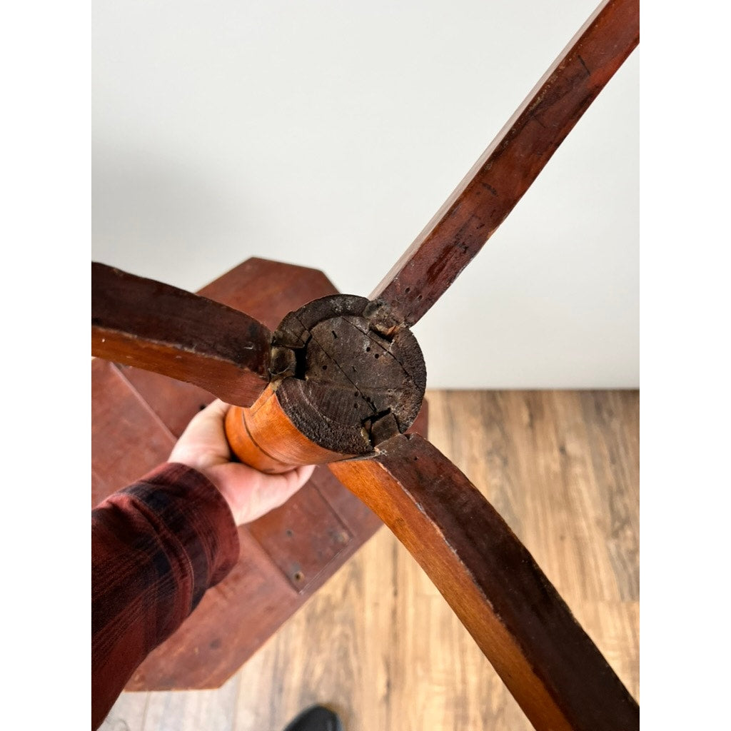 A hand holds the base of an Antique 19th Century Cut Corner Hepplewhite Candle Stand with three legs and visible cracks, set above a wooden floor against a white wall. Brand is unknown.