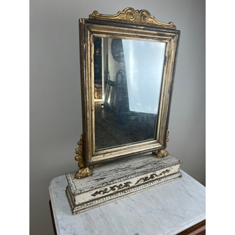 An Antique French Empire Gilt Wood Vanity or Table Mirror (c. Mid 19th Century) with an ornate gold frame and decorative base sits on a white marble surface, reflecting part of the room and blurred background objects.