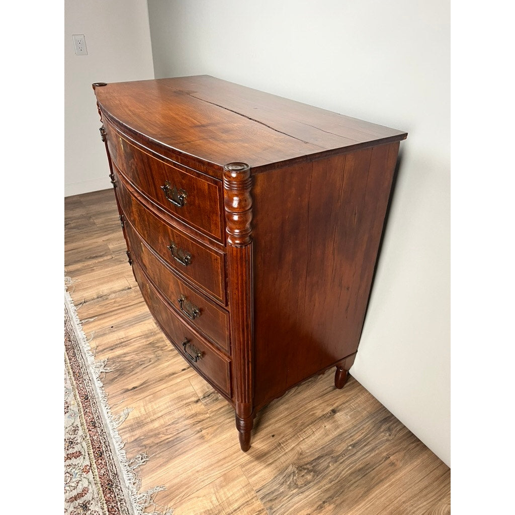 An Antique Sheraton Mahogany Bowfront Chest c. Early 19th Century with four curved drawers, ornate handles, and decorative side columns stands on a wood floor beside a white wall and patterned rug.