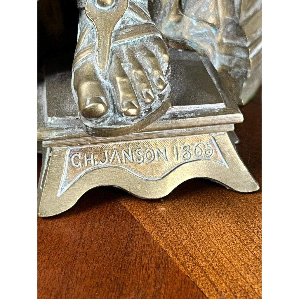 Close-up of the base of the antique bronze sculpture “Teacher and Pupil” by Louis Charles Janson (1865), signed Ch. Janson, showing a detailed sandal-clad foot. The piece rests on a wooden surface. Brand: None/Unknown.