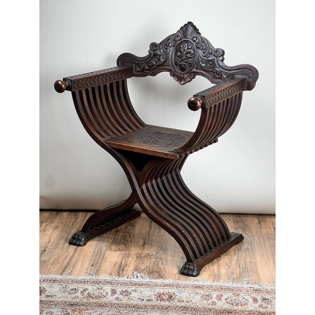 Antique Italian Renaissance Walnut Savonarola Chair, late 19th century, features curved slatted legs, carved floral details, rounded armrests, and paw feet—an elegant folding chair displayed on a wooden floor by a patterned rug.