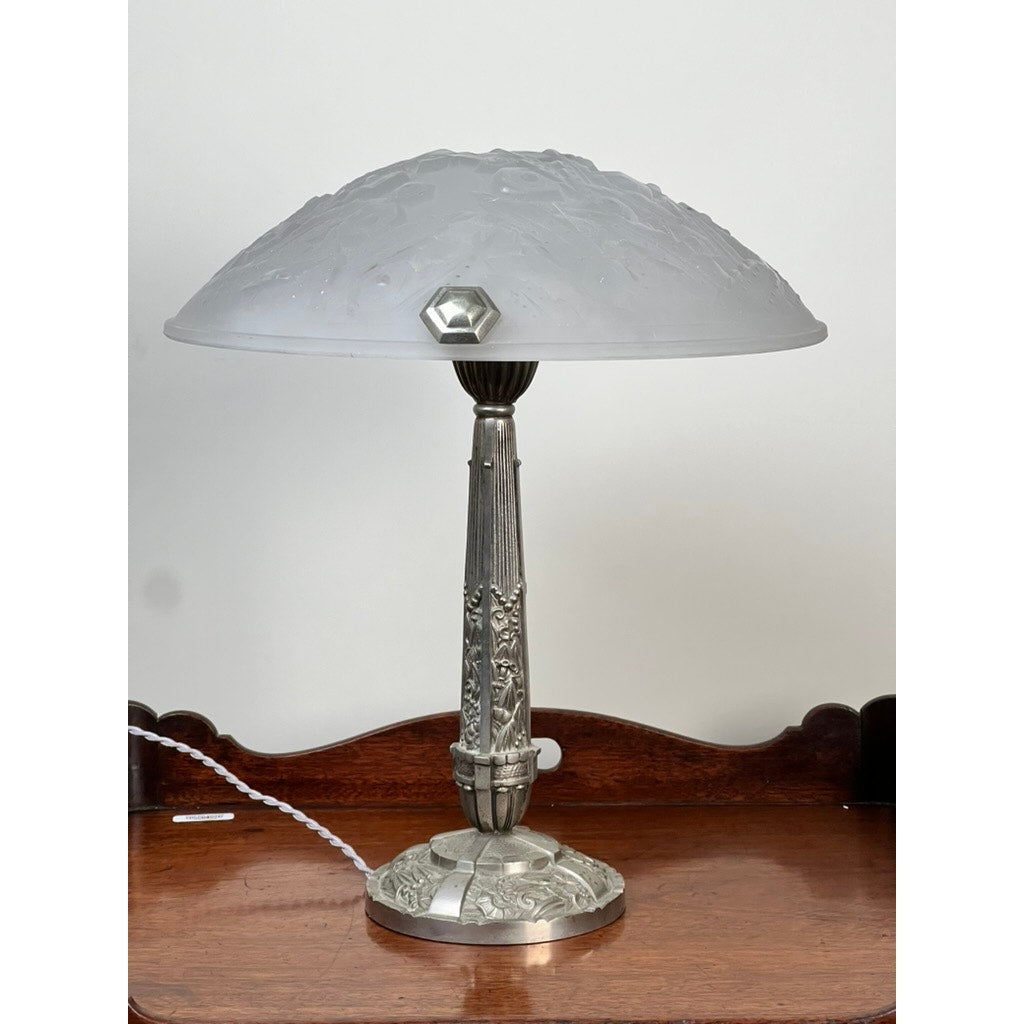 A Vintage French Art Deco Frosted Glass Table Lamp by Muller Frères Lunéville c. 1920, featuring ornate details and a frosted shade, sits on a polished wooden surface against a white wall with its white cord visible.