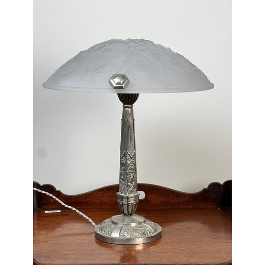 Vintage French Art Deco Frosted Glass Table Lamp by Muller Frères Lunéville c. 1920, offered by Pearl City Antiques, featuring a frosted glass shade and an ornate silver base. The lamp is displayed on a wooden surface against a plain white wall, with its white power cord visible on the left.