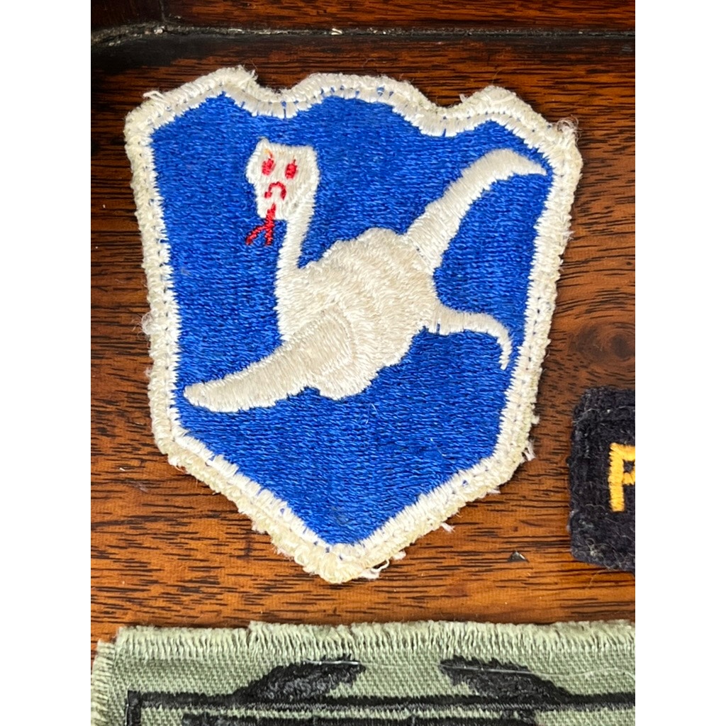 An embroidered patch with a white goose on a blue shield, red eyes, and open beak honors the WWII 158th Infantry Bushmasters—part of Frank W. Slatcher’s Pacific Theater grouping—sewn onto wood.