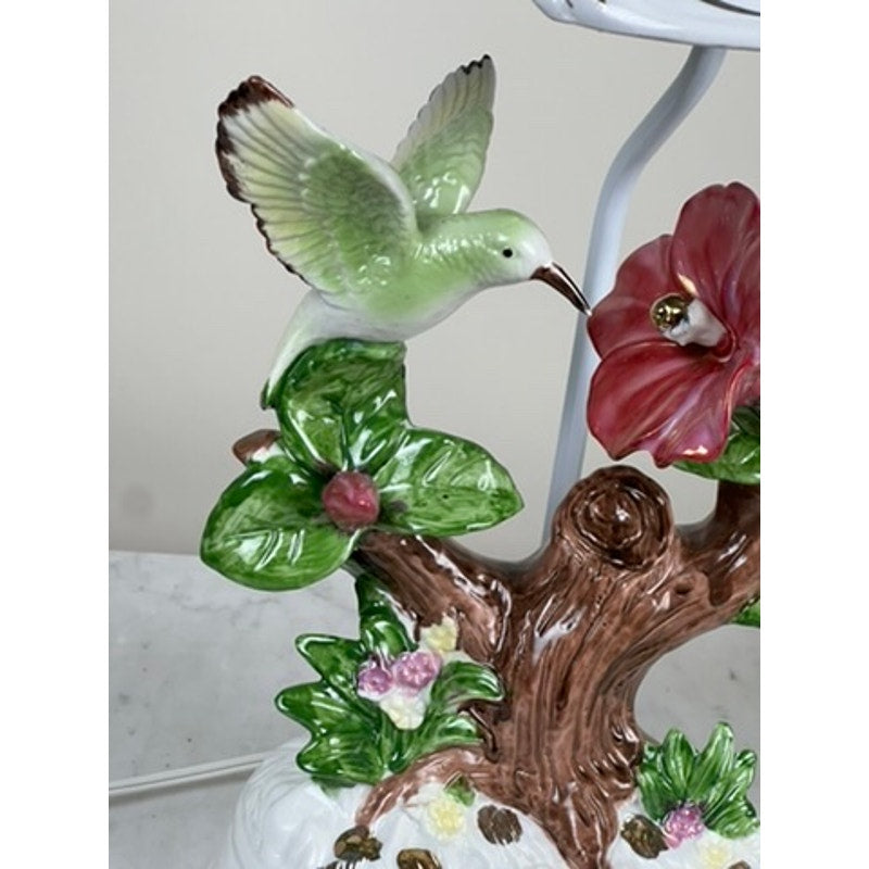 The Vintage Lena Liu Porcelain Hummingbird Accent Lamp depicts a green hummingbird hovering near a pink flower, perched on a tree branch with green leaves and colorful blossoms at the base. Brand is unknown.