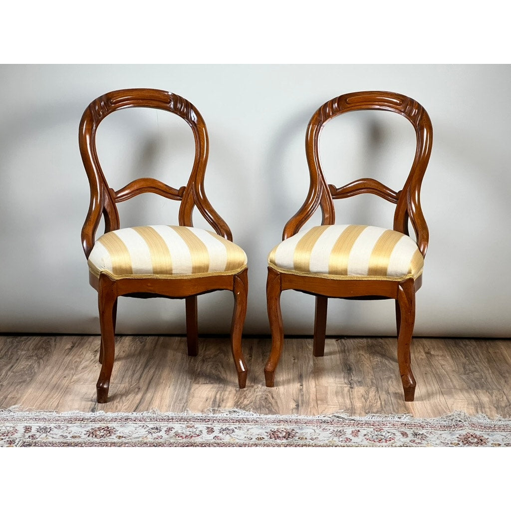 A Pair Antique Victorian Balloon Back Chairs c. Late 19th Century, featuring curved backs and yellow-and-white striped upholstered seats, sit side by side on a wooden floor with a patterned rug partially visible below.