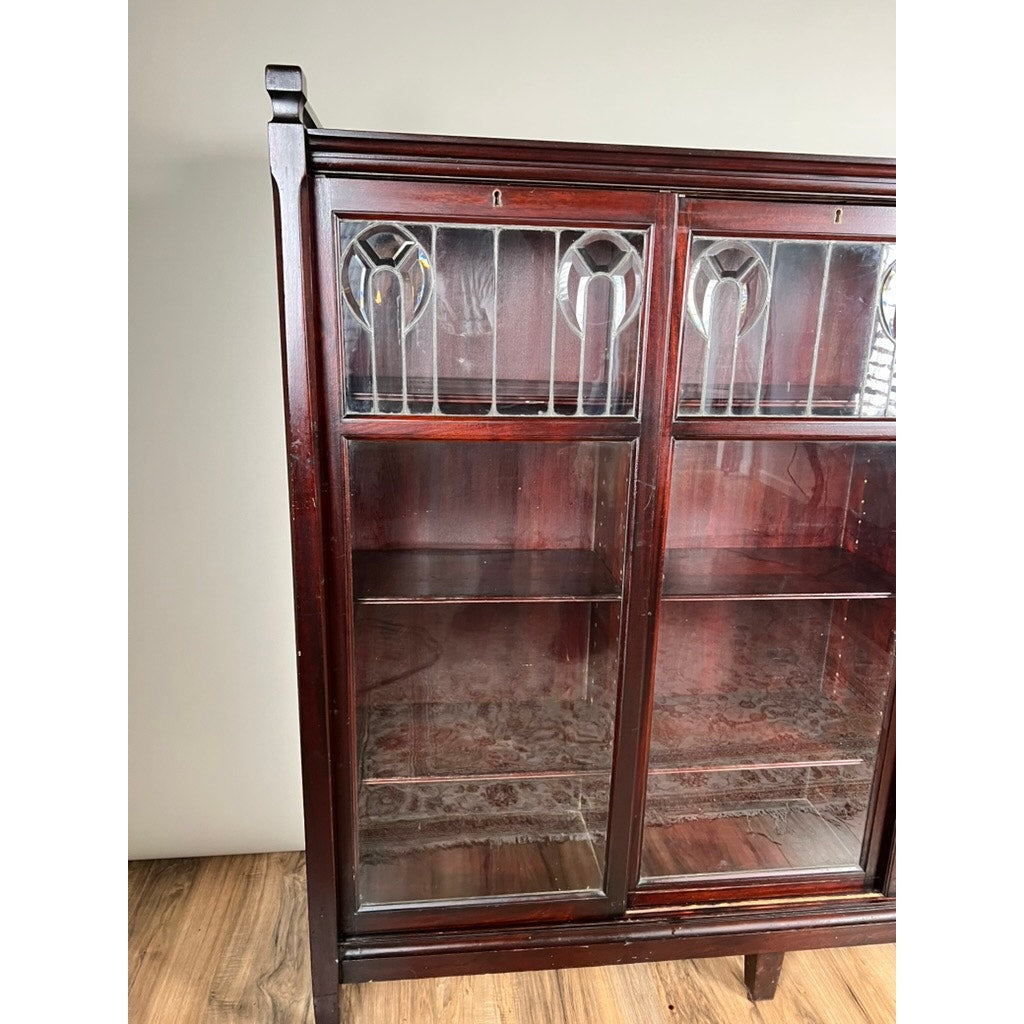 The Antique Arts & Crafts Leaded Glass Mahogany Bookcase c. 1920 features decorative leaded glass doors, empty shelves, and stands on a wooden floor against a plain light-colored wall. Brand: None/Unknown.