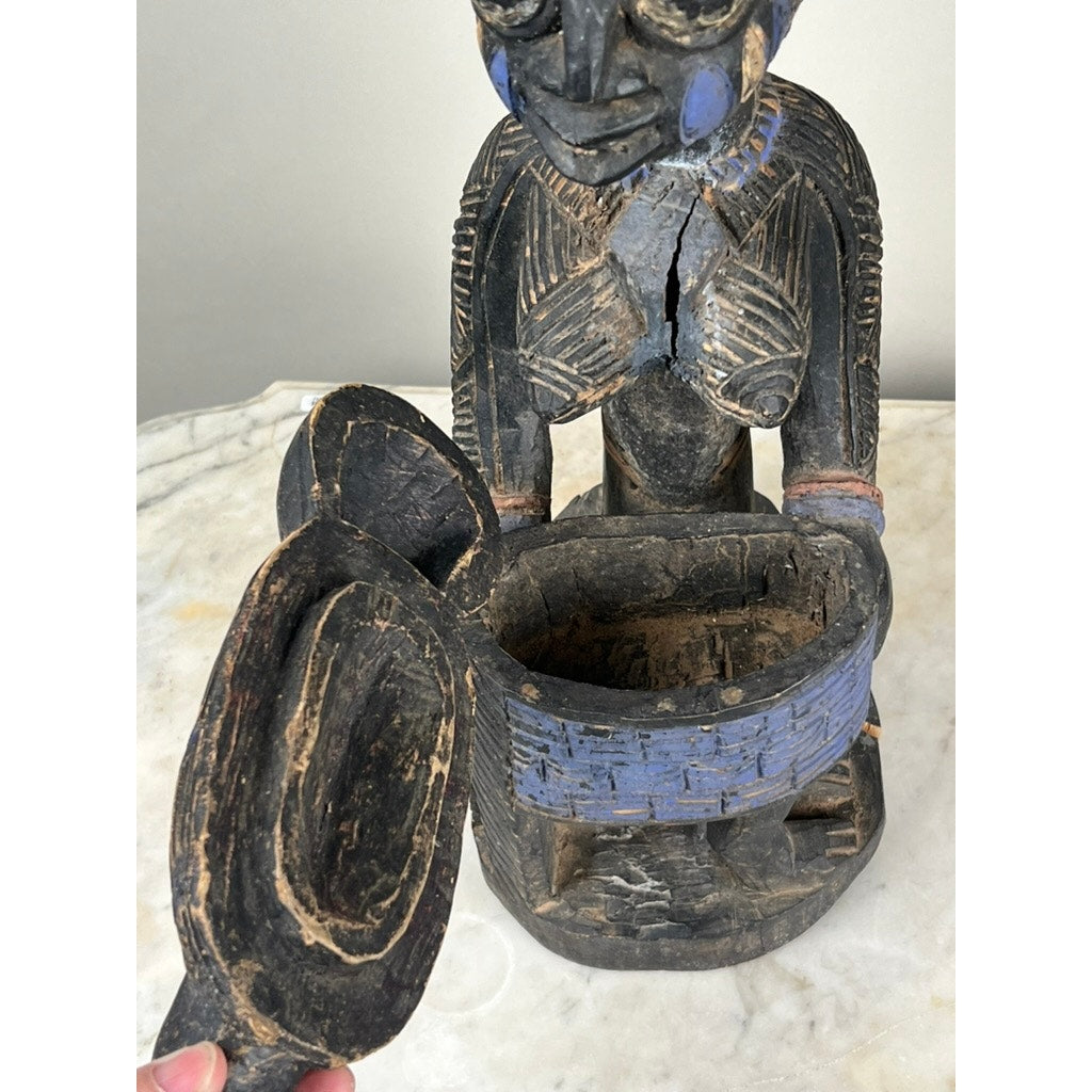 The Antique Igbo Ikenga Shrine Figure, probably from Nigeria, is a carved wooden piece with blue accents and a lidded bowl held at the waist. Lifting the lid reveals its hollow interior as it sits elegantly on a marble surface. Brand unknown.