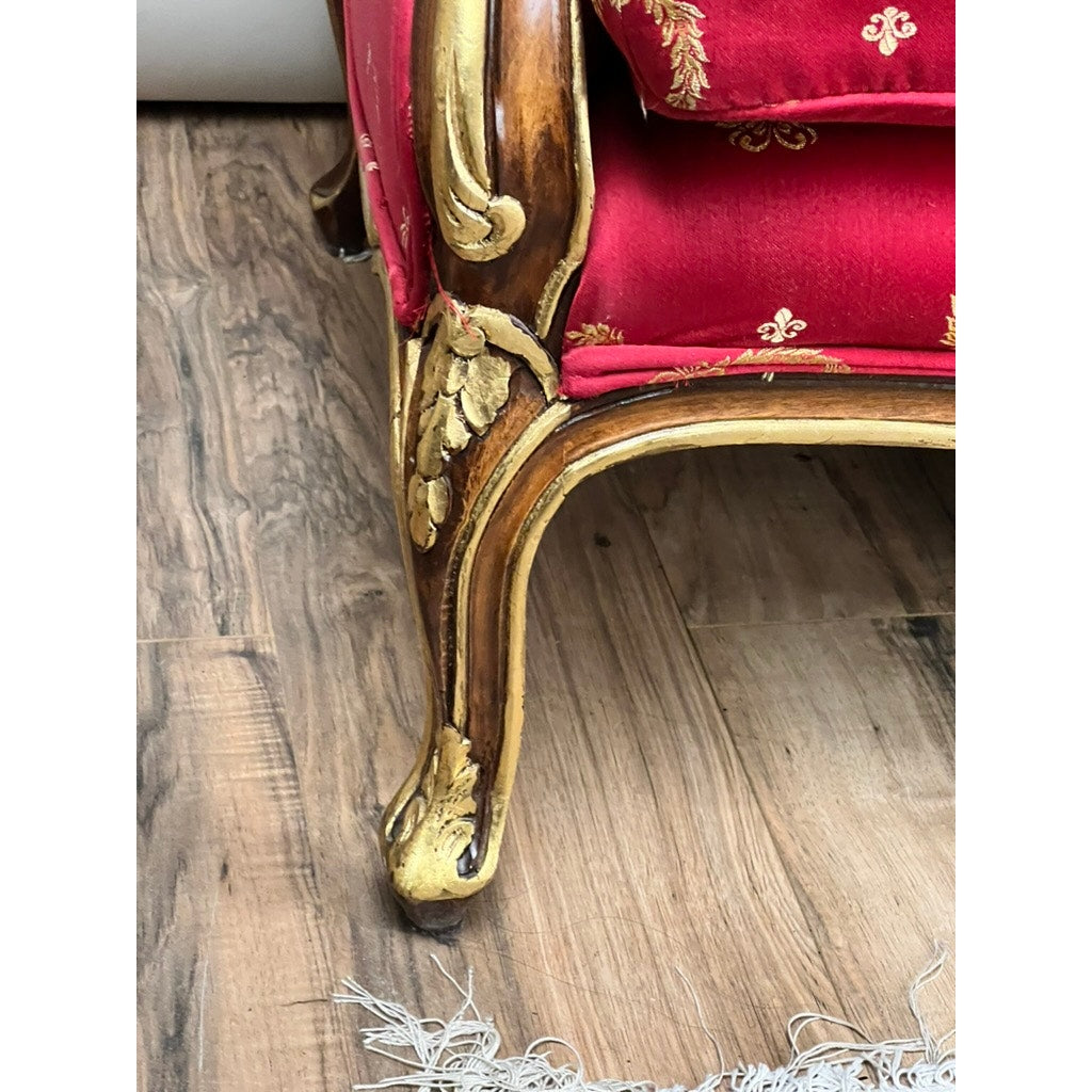 Detailed giltwood frame and wooden leg of a Vintage French Regency Style Custom Upholstered Settee with red fabric and gold accents on a wood floor. A white fringed rug appears partially in the lower right corner. Brand: None/Unknown.