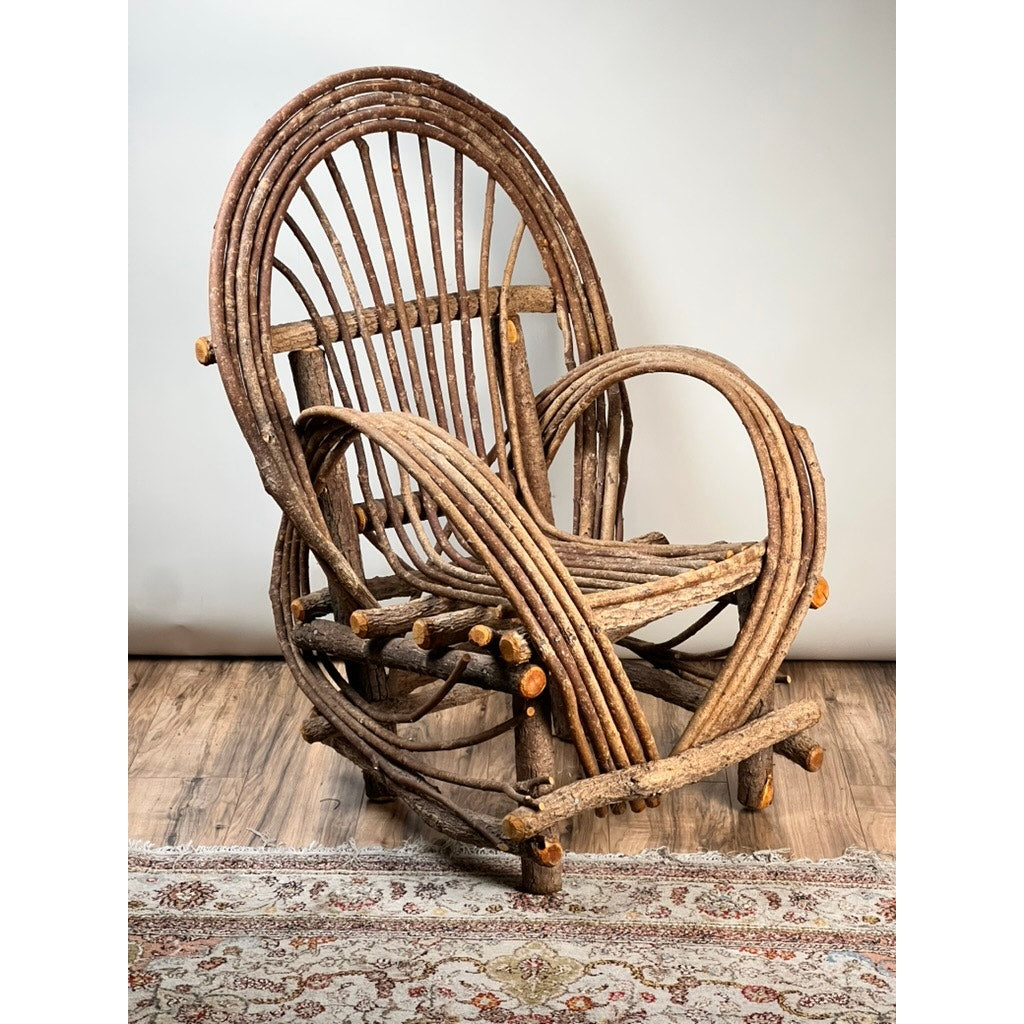 A Vintage Adirondack Bent Willow Arm Chair with curved, interwoven branches forms the back, arms, and legs. It sits on a wooden floor beside a patterned rug against a plain light-colored wall. Brand: None and/or Unknown.
